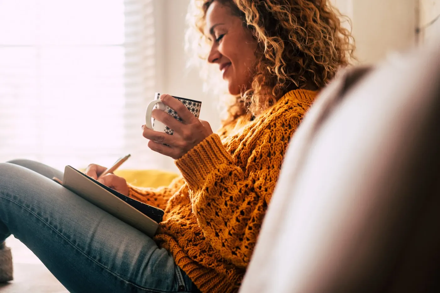 A woman curled up with a mug of coffee, writing in her journal